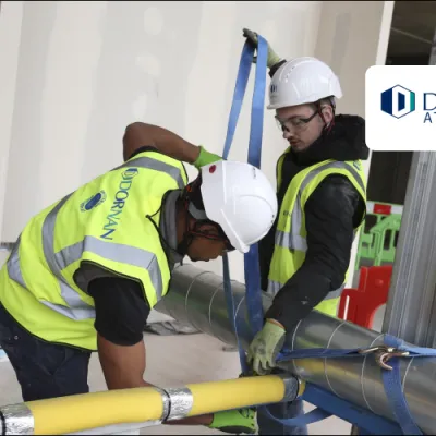 Two construction workers in high vis jackets, white hard hats, and goggles work on tying a pipe