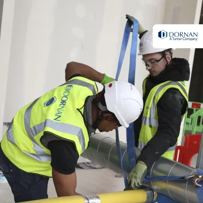 Two construction workers in high vis jackets, white hard hats, and goggles work on tying a pipe