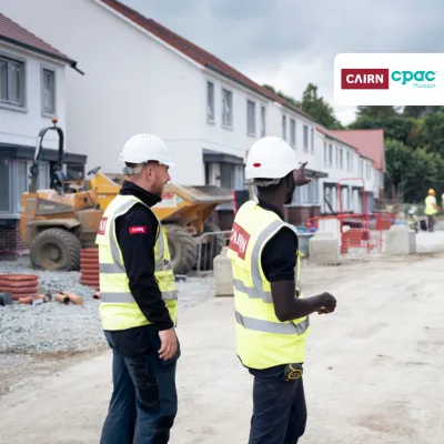 Three construction professionals in safety gear and hard hats are standing on a residential building site, discussing progress. Houses under construction are visible in the background.