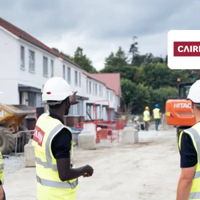 Three construction professionals in safety gear and hard hats are standing on a residential building site, discussing progress. Houses under construction are visible in the background.