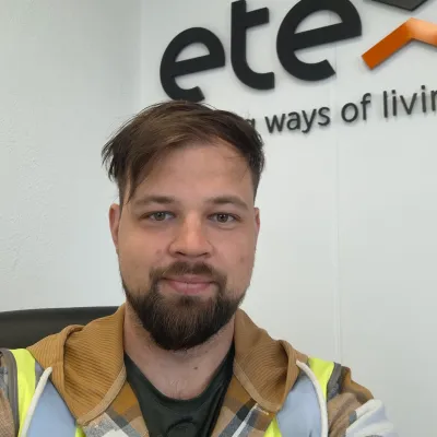 A person wearing a high-visibility vest and a hoodie, smiling in front of a white wall with the logo "etex"  visible in the background.