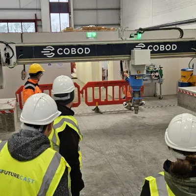 Group of people in hard hats and high-vis vests observing a 3D concrete printer inside an industrial facility.
