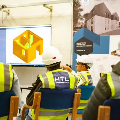 Construction workers in HTL vests and hard hats attend a classroom training session with a 3D model displayed on a large screen.