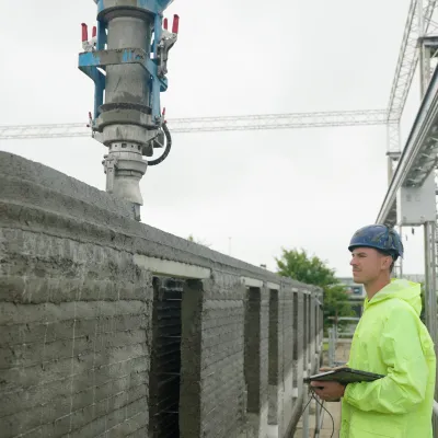 A construction worker in a hard hat and high-vis jacket observes a 3D printer building a concrete wall.