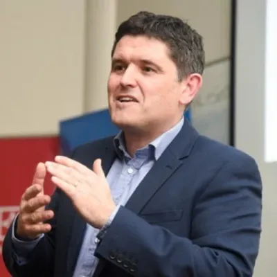 A person in a suit speaking in front of a presentation screen