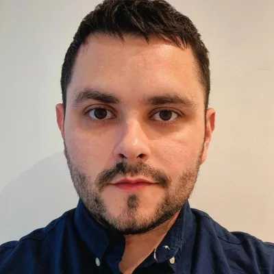 A person with short dark hair and beard, wearing a dark collared shirt, standing in front of a plain light wall.