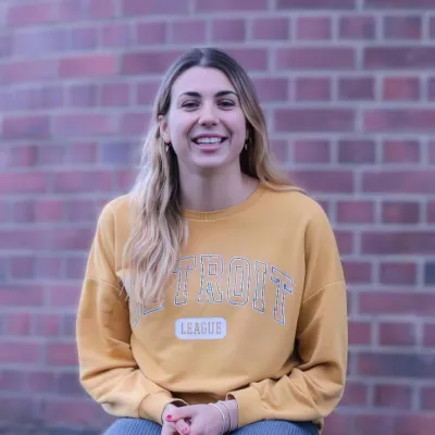 Person sitting on a low brick wall, smiling, wearing a yellow sweatshirt and jeans