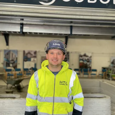 A person wearing a yellow high-visibility jacket standing indoors in front of a sign that reads "COBOT" and various 3d printing equipment in the background.