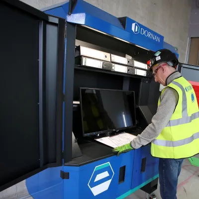 A construction worker in a hard hat reviews notes on a clipboard in front of a computer