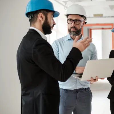 Three engineers wearing formal clothing and safety helmets discussing a construction project indoors, with one holding a laptop.