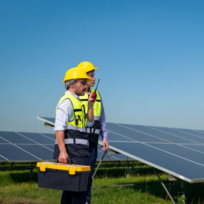 A construction worker wearing a yellow hard hat and safety vest stands beside a solar panel array, holding a toolbox in a sunny outdoor environment.