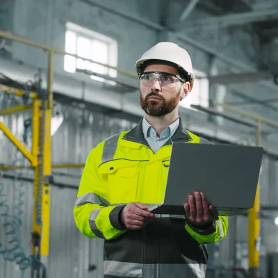 Person in a hard hat and high-visibility jacket using a laptop at an industrial facility.