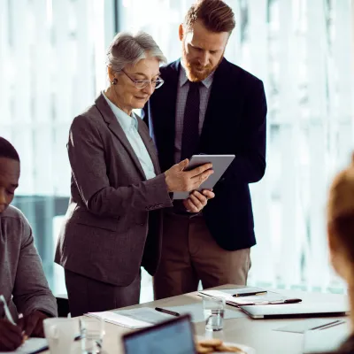 Group of professionals in a modern office having a discussion around a table, with one person showing a tablet to a colleague.