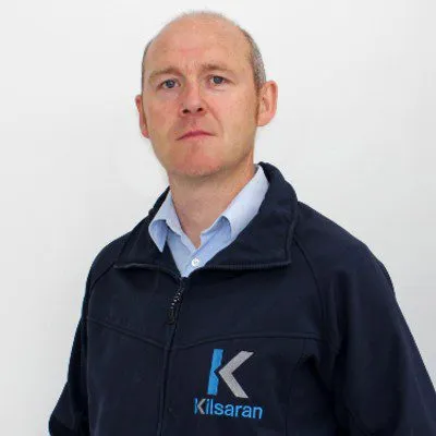 Person wearing a dark Kilsaran jacket with a light blue shirt underneath, standing against a plain white background and looking at the camera.