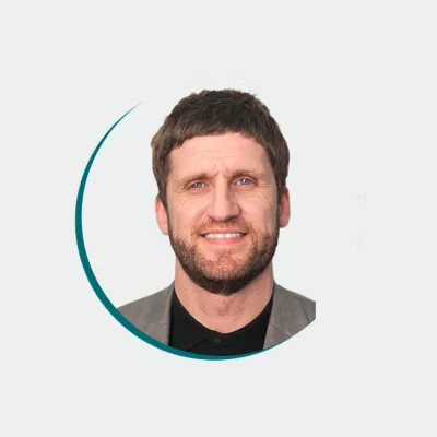 a person wearing a jacket over a dark shirt, smiling at the camera against a light background.