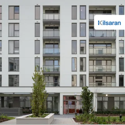 Front view of a modern apartment building with several balconies, large windows, and a landscaped entrance area with plants and trees.