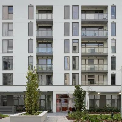 Front view of a modern apartment building with several balconies, large windows, and a landscaped entrance area with plants and trees.