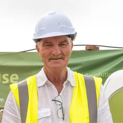 Person wearing a white hard hat, yellow high-visibility vest, and white shirt with glasses hanging from the collar, standing outdoors in front of a green banner.