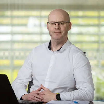 Person wearing glasses and a white shirt sitting at a desk with a laptop and notepad, looking at the camera with hands folded.