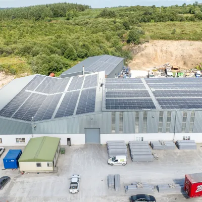 Industrial facility with large solar panels on the roof, surrounded by parked vehicles and storage materials.