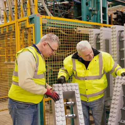 Two people in high-visibility jackets inspecting large grey insulation blocks inside a manufacturing facility, with machinery and safety fencing in the background.