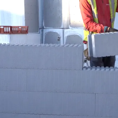 Construction worker in a high-visibility vest placing a large grey insulation block onto a wall under construction, with stacked blocks and materials in the background.
