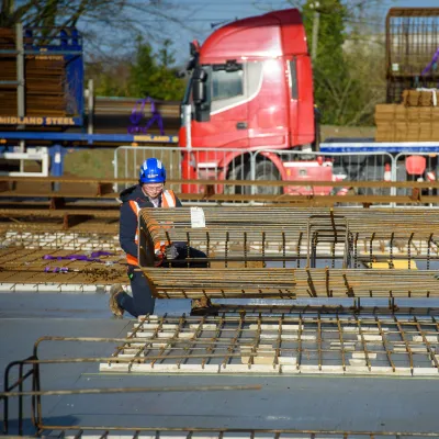 Construction worker in safety gear handling steel reinforcement bars on a site, with a red truck and stacked rebar in the background.