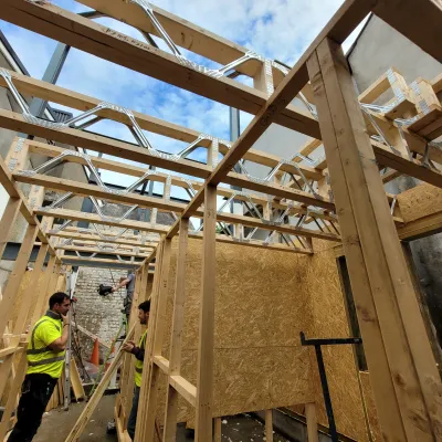 Construction workers in high-visibility vests assembling a timber frame structure with exposed beams and panels on a building site.