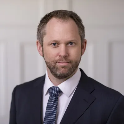 Person in a dark suit and tie facing the camera with a neutral expression, standing in front of a light-colored wall.