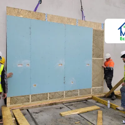 Workers in safety gear installing a large prefabricated wall panel on a construction site, with MMC Eco Homes logo in the corner.