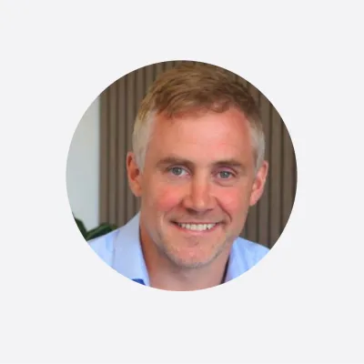 Portrait photo of a smiling man with short light hair, wearing a light blue shirt, against a neutral background