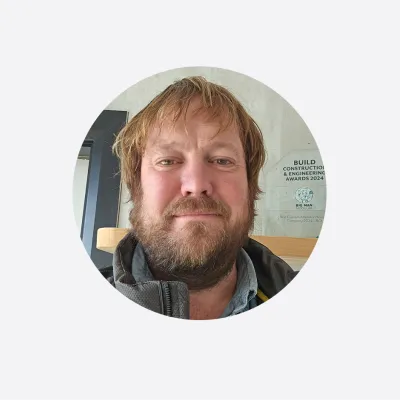 Portrait photo of a man with light brown hair and beard, wearing a jacket, with an award plaque visible in the background.