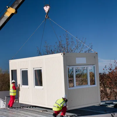 Prefabricated modular unit being lifted by a crane onto a site, with two workers in safety gear guiding it into place.