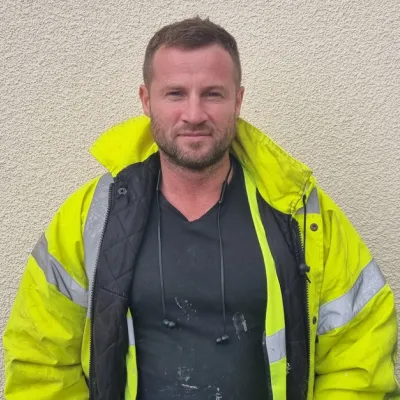Construction worker wearing a high-visibility yellow jacket and black shirt standing against a light-coloured wall.