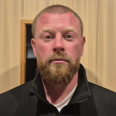 Portrait of a person with a short beard and close-cropped hair, wearing a black jacket, standing against a wooden wall.