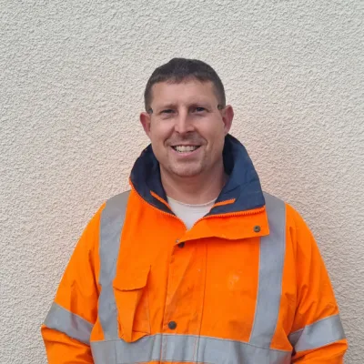 Portrait of a person wearing a bright orange high-visibility jacket, standing against a light-coloured wall and smiling at the camera.