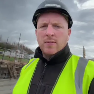 Person wearing a high-visibility vest and safety helmet on a construction site, with scaffolding and cloudy sky in the background.