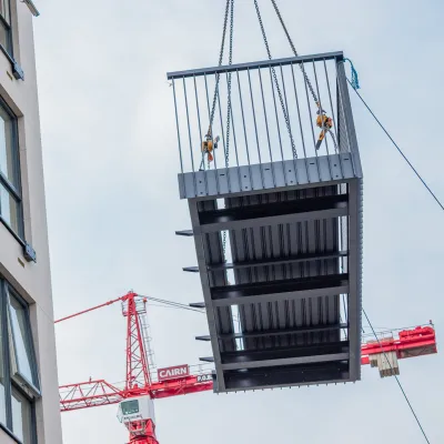 Crane lifting a prefabricated metal balcony module into place beside a building.