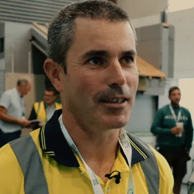 Portrait of a person wearing a yellow construction polo shirt and high-visibility vest, with a lanyard around their neck, standing indoors at an event with other people in the background.