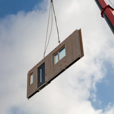 Crane lifting a prefabricated wall panel with a door and windows against a cloudy sky.