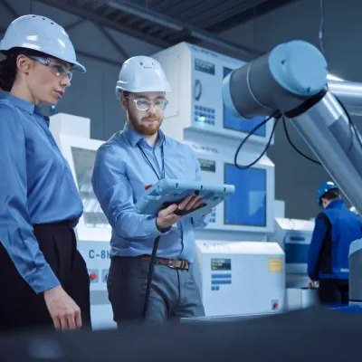 Two engineers in hard hats use a tablet to program a robotic arm on a factory floor.