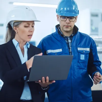 Two people wearing hard hats in an industrial setting, one in a business suit holding a laptop and the other in a blue work jacket, reviewing information together.
