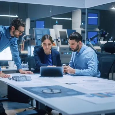 Three people in a modern office sitting around a table with a laptop, reviewing documents and technical drawings, with machinery and monitors visible in the background.