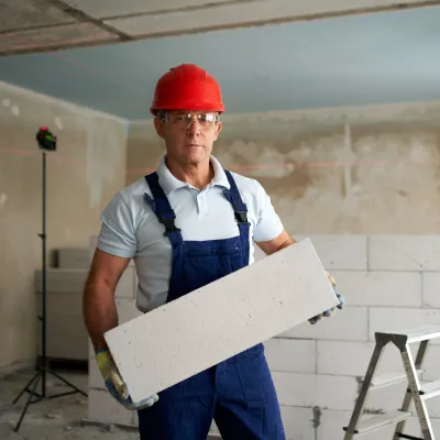 Construction worker in a red hard hat and overalls holding a large white block indoors, with a step ladder and unfinished wall in the background.