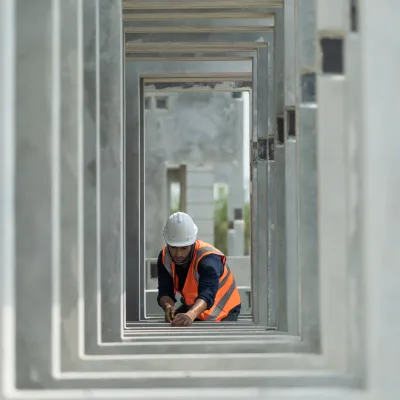"Person wearing a hard hat and orange safety vest working with a measuring tool inside a series of concrete rectangular frames at a construction site.