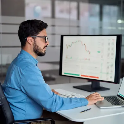 Person in a blue shirt working at a desk with a laptop and large monitor displaying financial charts and data.