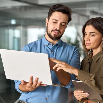 Two people standing in an office, holding a laptop and a tablet, discussing something on the laptop screen.