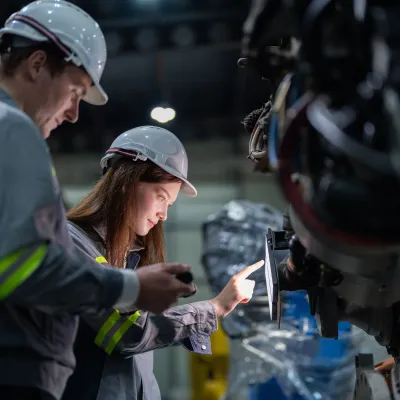 Two people wearing safety helmets and reflective clothing inspecting industrial machinery, with one person pointing at a control panel.