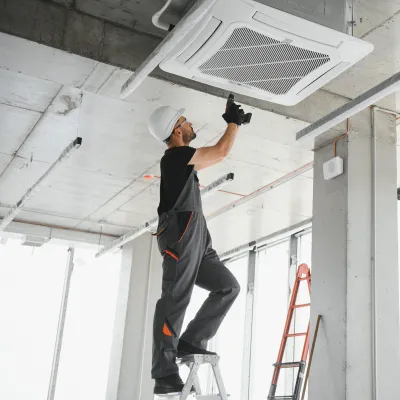 Person wearing a hard hat and overalls standing on a ladder and installing or servicing a ceiling-mounted air conditioning unit in a modern building.