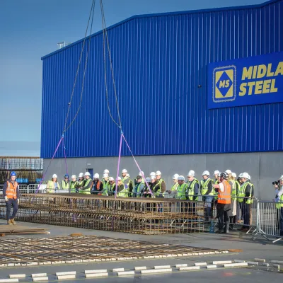 A group of workers in safety vests gathered outside a large blue Midland Steel facility building.